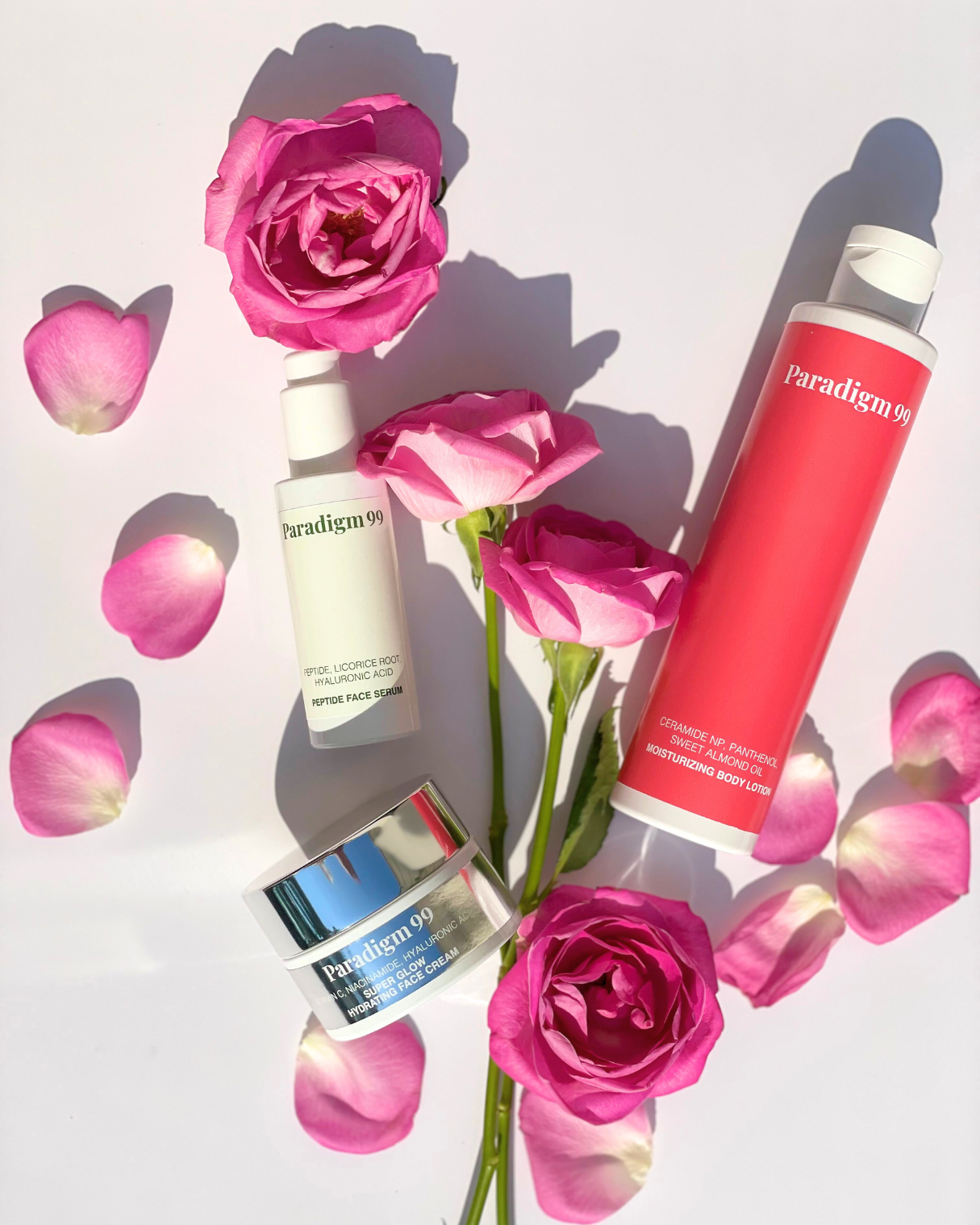 Paradigm skincare products with pink roses on a white background. Products are Peptide face Cream, Moisturizing body cream with ceramides, sweet almond oil, panthenol and super glow face cream with vitamin c, hyaluronic acid, niacinamide. It is healthy glow ritual. 