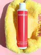 Body lotion bottle labeled 'Paradigm 99' on a yellow textured surface with pink background.