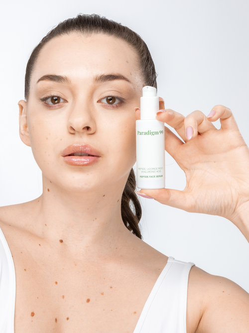 A woman with brown hair in a ponytail holding a paradigm99 peptide serum in a light green glass bottle with a dispenser. She has beautiful, well cared for skin with a few moles on her right cheek.