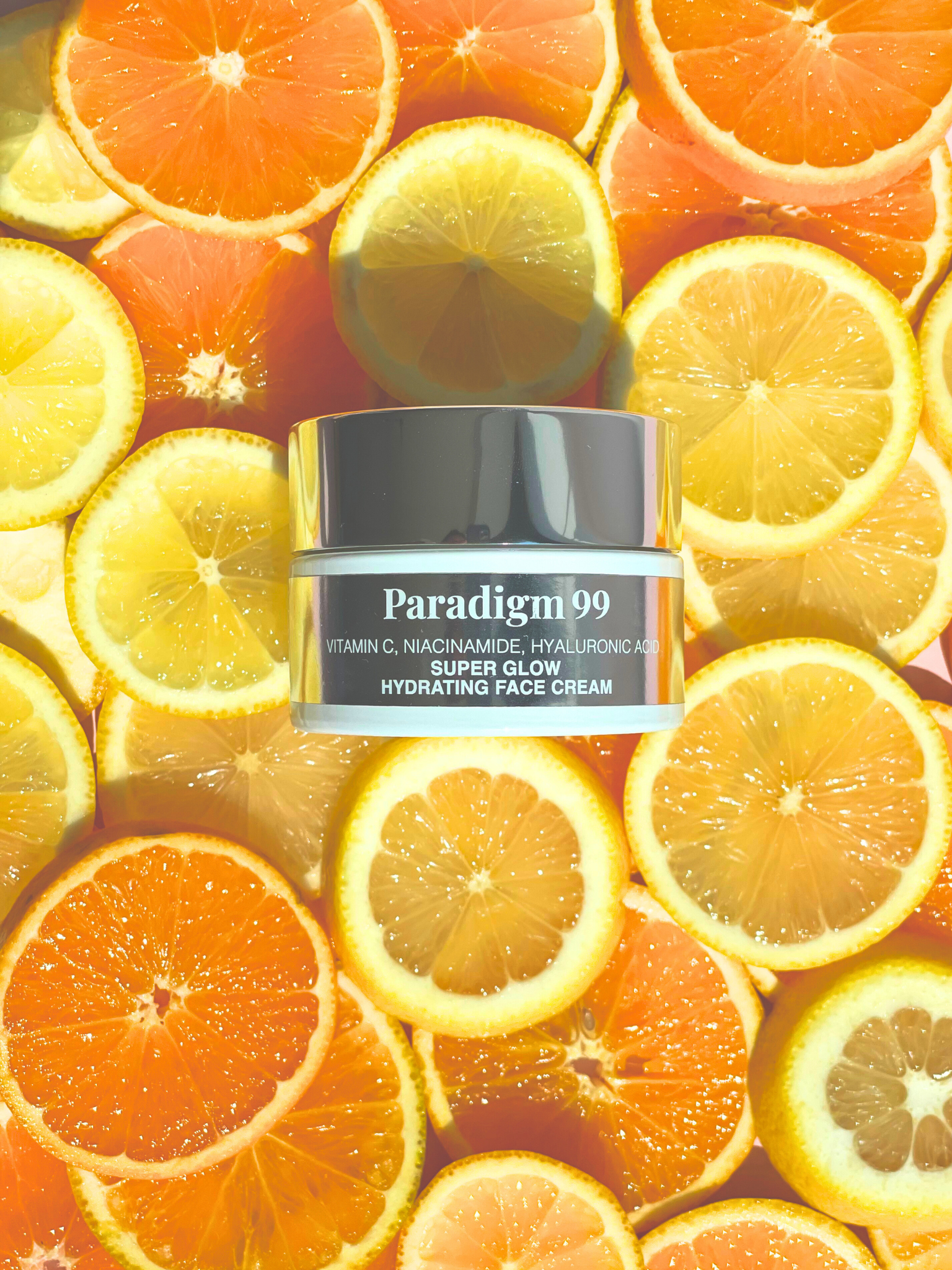 Paradigm 99 Super Glow Face Cream contains Vitamin C, Niacinamide, Hyaluronic Acid and glass jar  surrounded by sliced oranges and lemons. Photo looks bright and fresh. 
