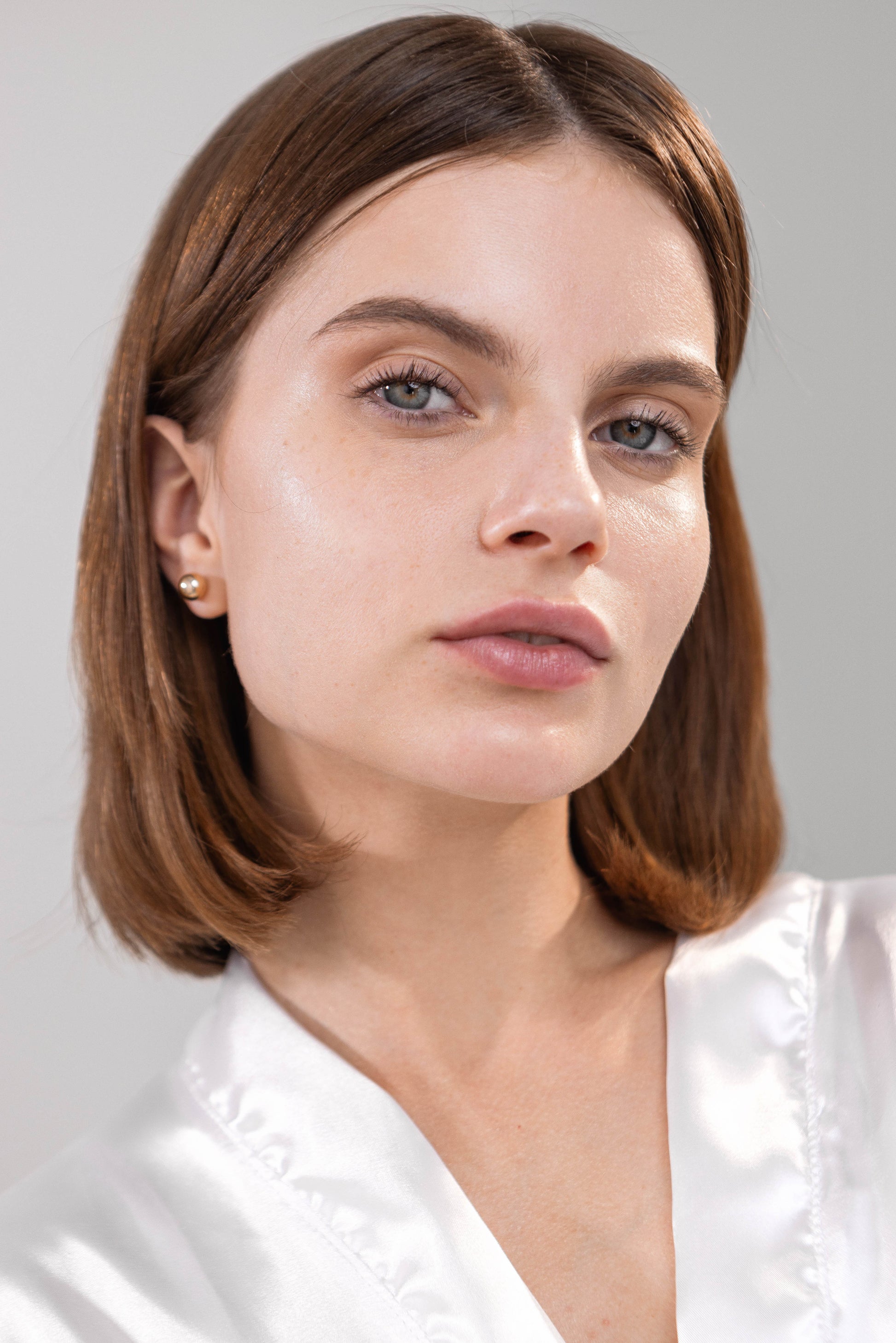 Woman with short brown hair wearing a white top against a light grey background with glow and fresh skin. 